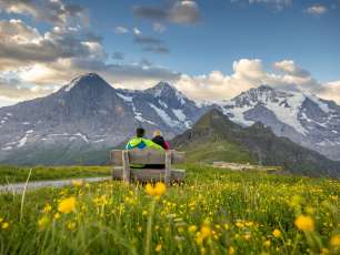Deux randonneurs sont assis sur un banc. À l'arrière-plan, l'Eiger, le Mönch et la Jungfrau ; au premier plan, une prairie fleurie jaune. 