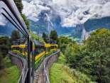 Die Wengernalpbahn fährt entlang einer Hangstrecke durch grünes Talgebiet. Im Hintergrund öffnet sich das Lauterbrunnental mit steilen Felswänden und markanten Gipfeln unter wechselhafter Bewölkung.