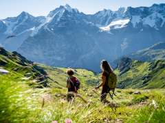 Zwei Freundinnen wandern im Blumental in Mürren. Sie geniessen den Ausblick über die grüne Landschaft mit dem weissen Bergpanorama mit Eiger, Mönch und Jungfrau im Hintergrund.